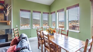 Un comedor con mesa y sillas de madera, un jarrón de flores y ventanas amplias con vistas a un paisaje montañoso.
