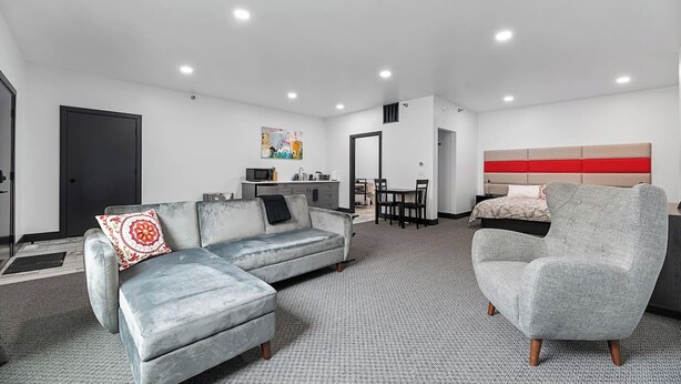 Ein modernes Hotelzimmer mit grauem Sofa, einem grauen Sessel, einem Essbereich und einem Bett mit roter Kopfteil.
