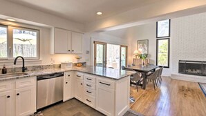 A modern kitchen with white cabinetry, a built-in dishwasher, and a large island with a sink. There is a dining area with a wooden table and chairs, and a fireplace in the background. - Upscale Hamptons Home: 1 Mi to Sammys Beach! (East Hampton)