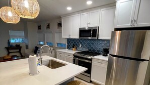 A modern kitchen with white cabinets, a stainless steel refrigerator, a microwave, and a stove. There are two wicker stools and a large island with a sink. - Kings Kamp (Key Largo)