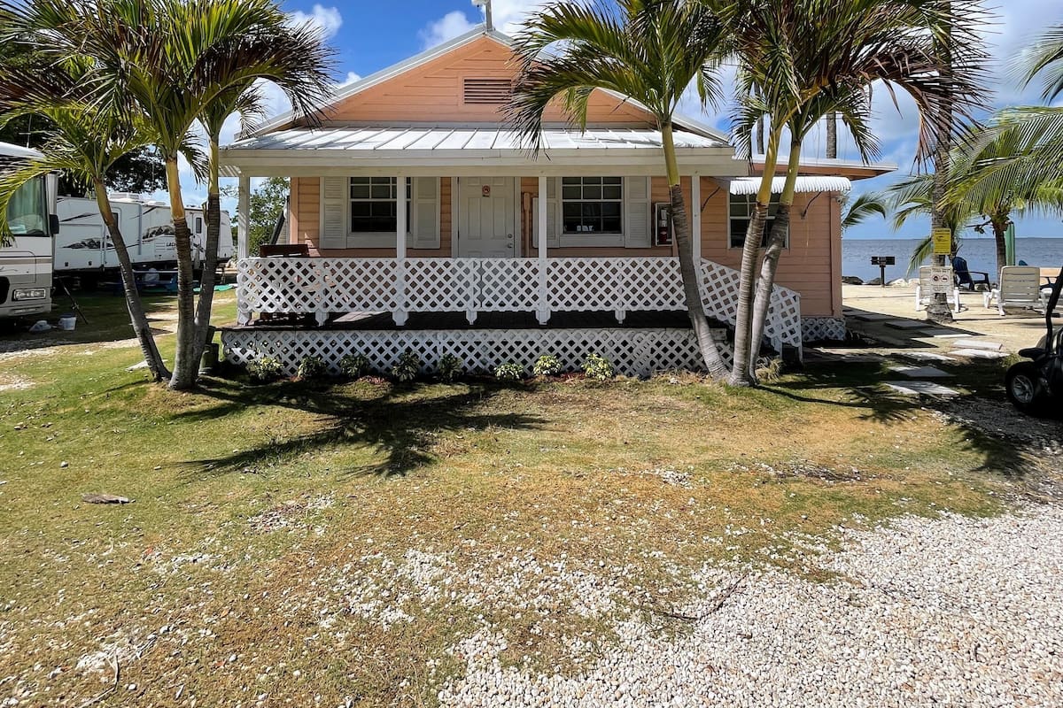 A single-story house with a porch, surrounded by palm trees and a gravel driveway.