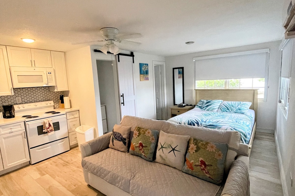 A compact living space with a kitchenette, a sofa with decorative pillows, a bed with a blue and white striped bedspread, and a ceiling fan.