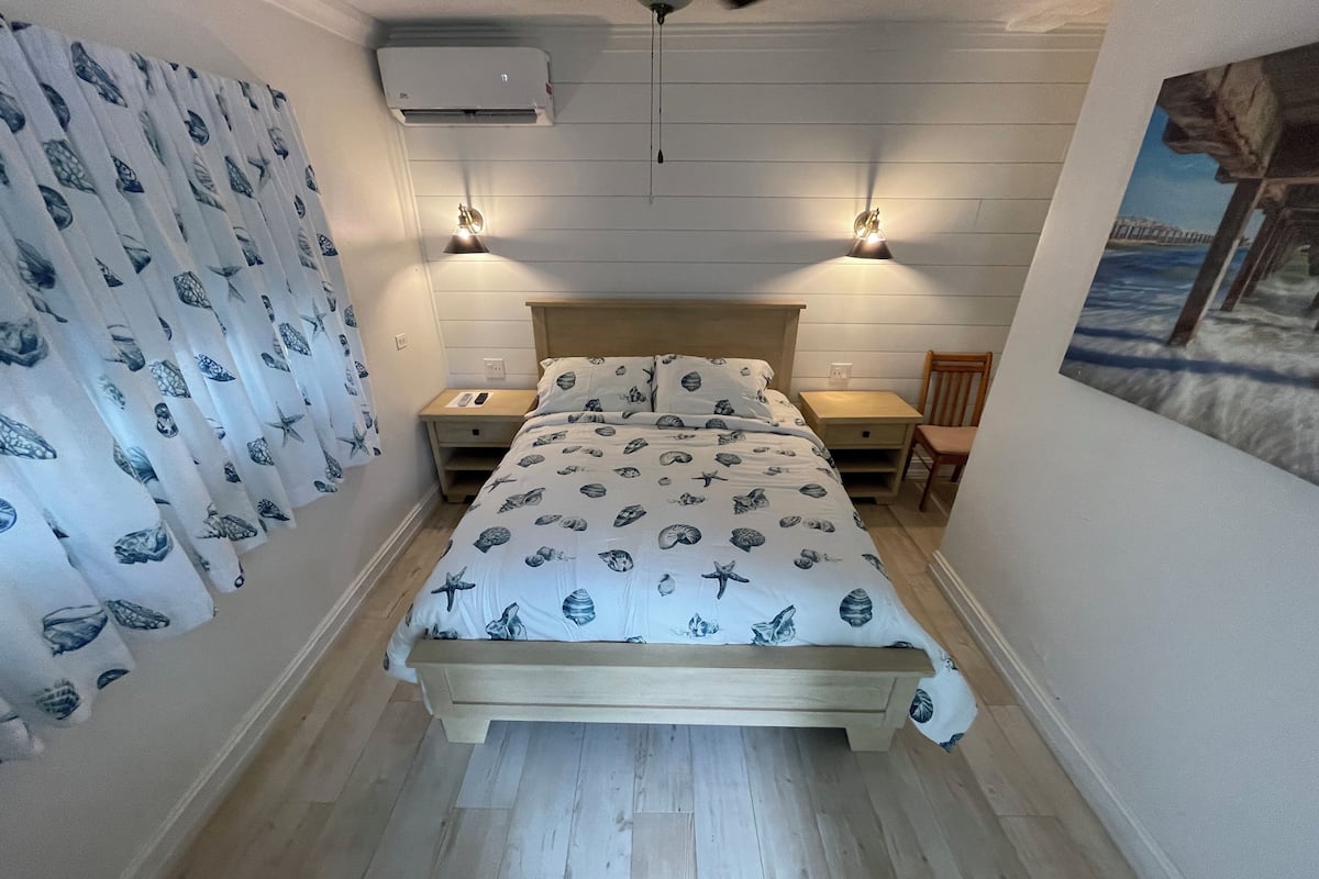 A bedroom with a bed, two bedside tables, a chair, a wall-mounted air conditioner, and a painting.
