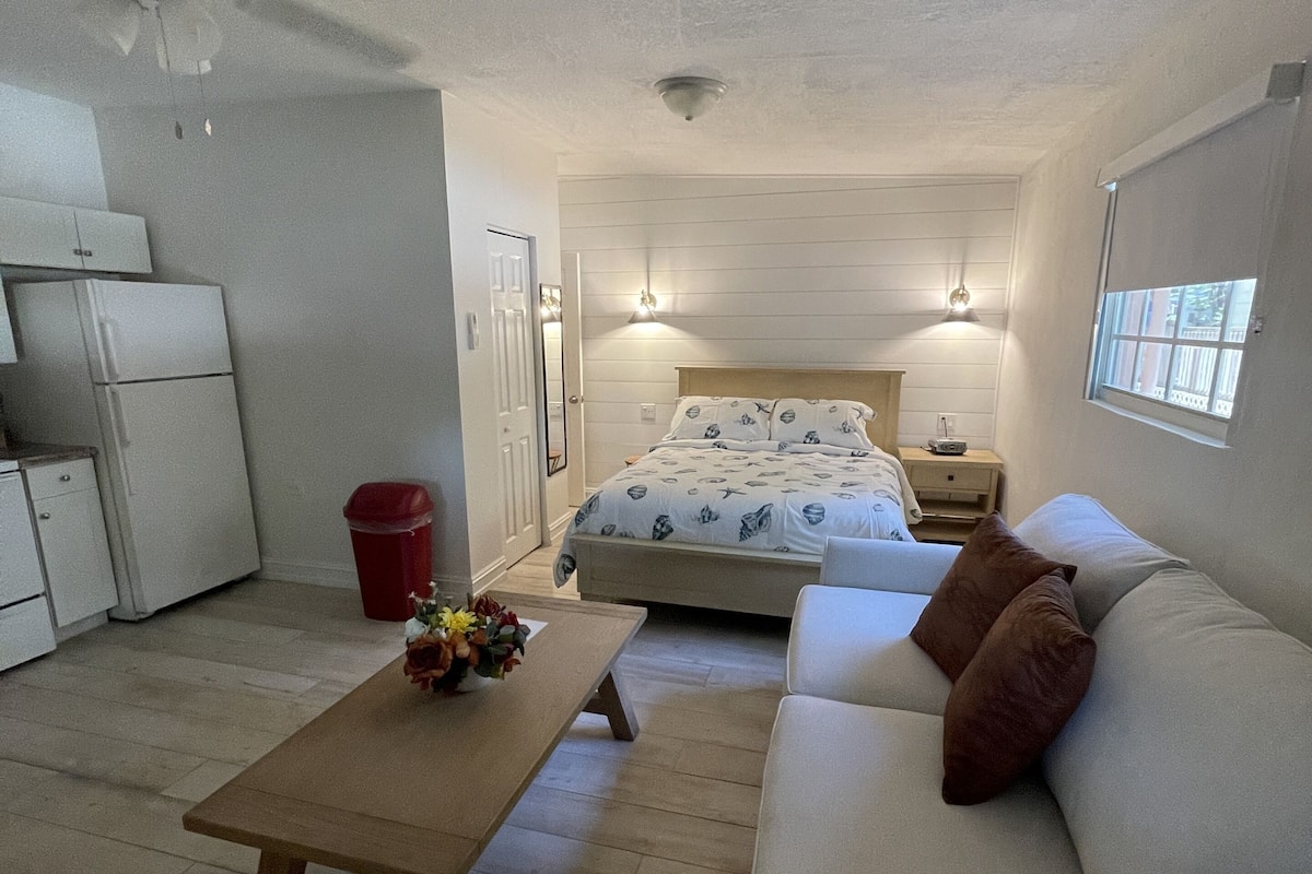 A compact room with a bed, a sofa, a small table, a ceiling fan, and a window.