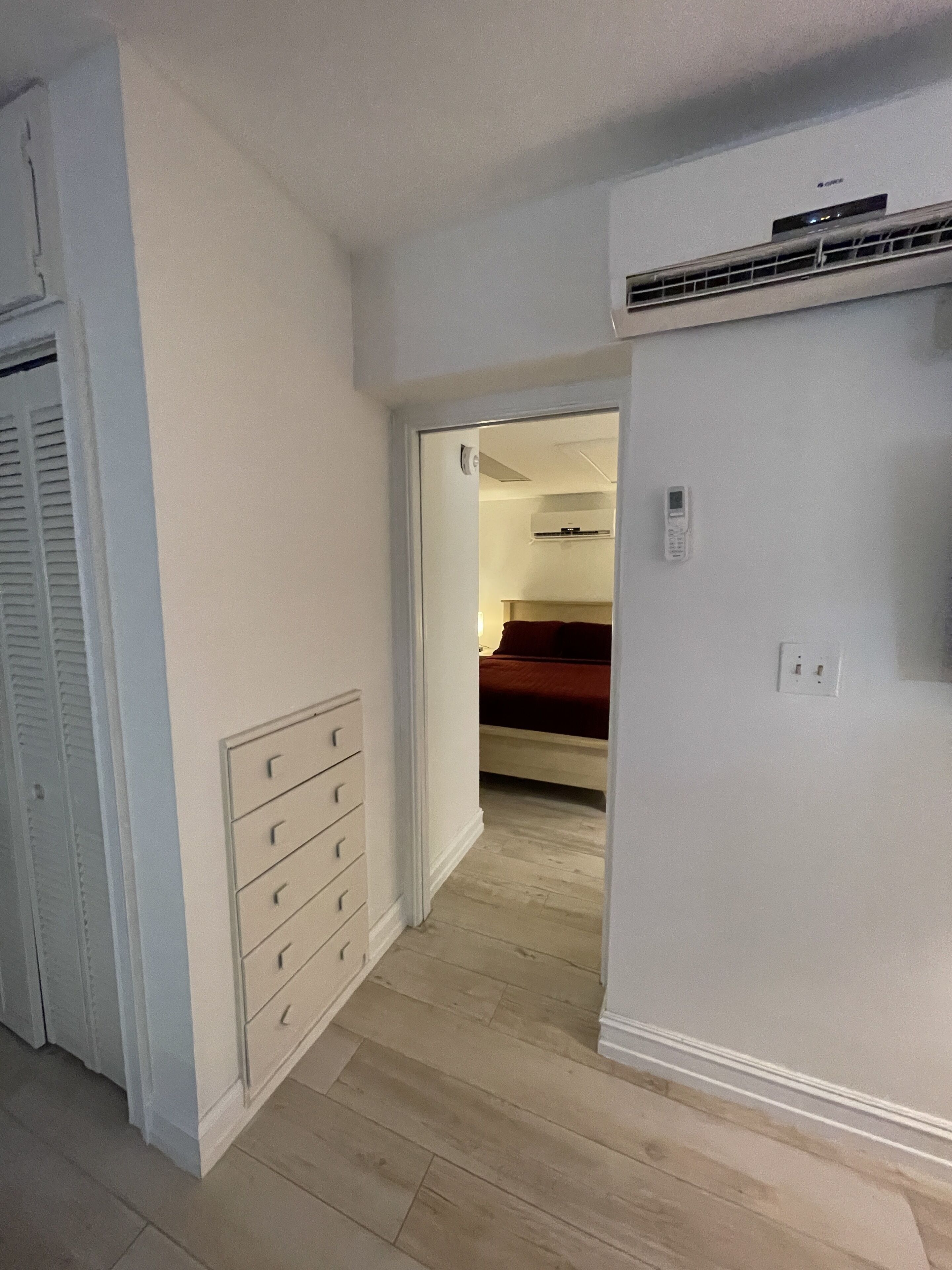A room with a wooden floor, a white wardrobe, a bed with a red blanket, and a white air conditioning unit on the wall.