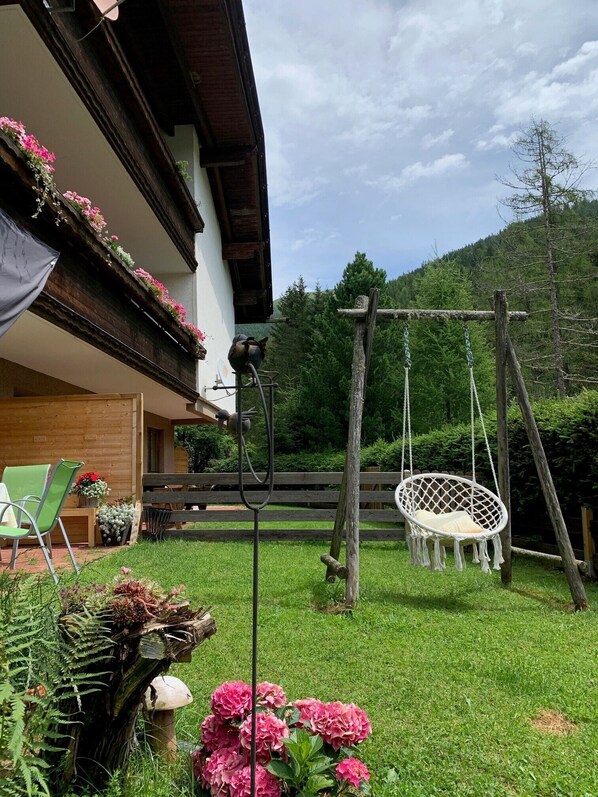 A rustic wooden building with a balcony adorned with flowers, a swing chair, and a hanging chair in a lush garden.