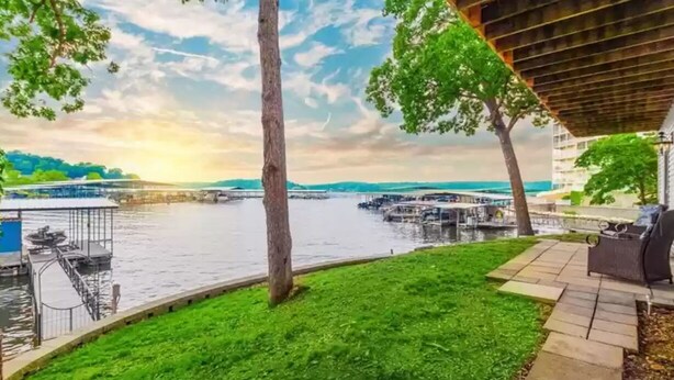 Un patio junto al río con césped, una zona cubierta para sentarse y vistas a un puente y al agua.