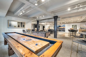 Une table de billard, une table de baby-foot et un coin bar dans une pièce spacieuse aux murs et au plafond blancs, éclairée par des luminaires.
