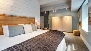 A modern bedroom with a large bed, a wooden headboard, a wardrobe, and a small round ottoman.