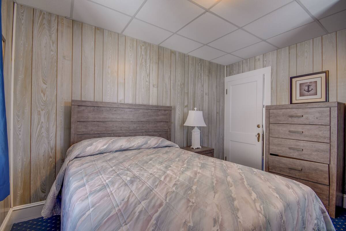 Un dormitorio con cabecera de madera, cama con colcha estampada, mesita de noche con lámpara, cómoda y una puerta con un cuadro enmarcado.