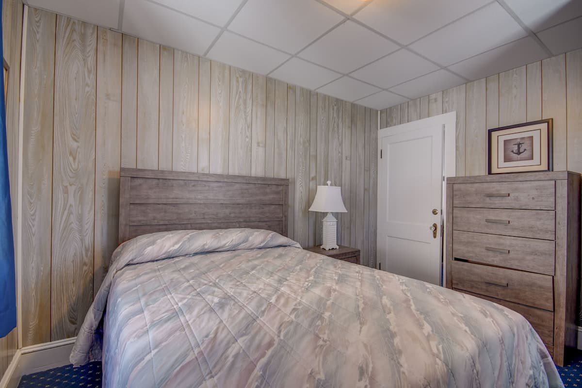Quarto com cabeceira de madeira, cama com colcha estampada, mesa de cabeceira com abajur, cômoda e porta com quadro.