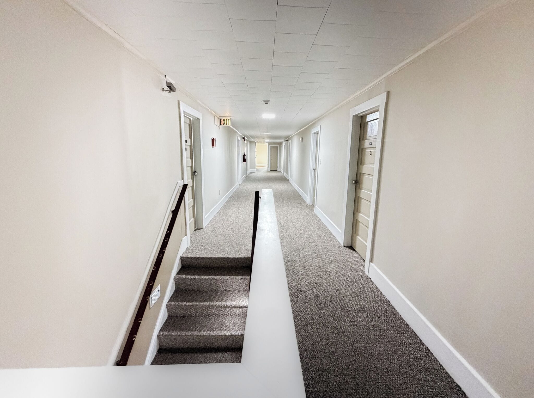 A long, narrow hallway with carpeted stairs leading to a door at the end.