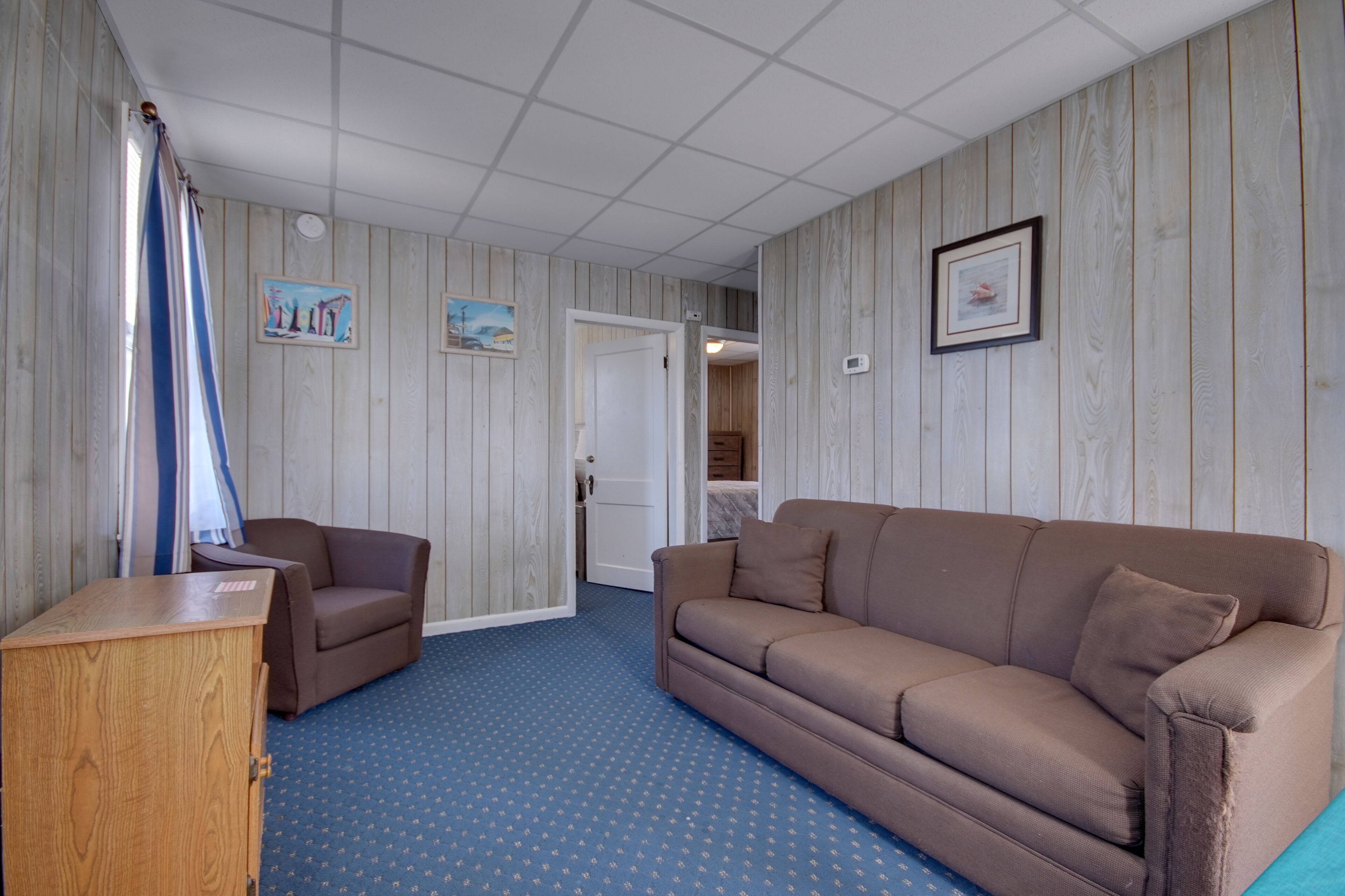A room with a brown sofa, a wooden cabinet, and a blue carpet. There are two paintings on the wall and a door leading to another room.