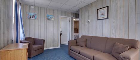 A room with a brown sofa, a wooden cabinet, and a blue carpet. There are two paintings on the wall and a door leading to another room.