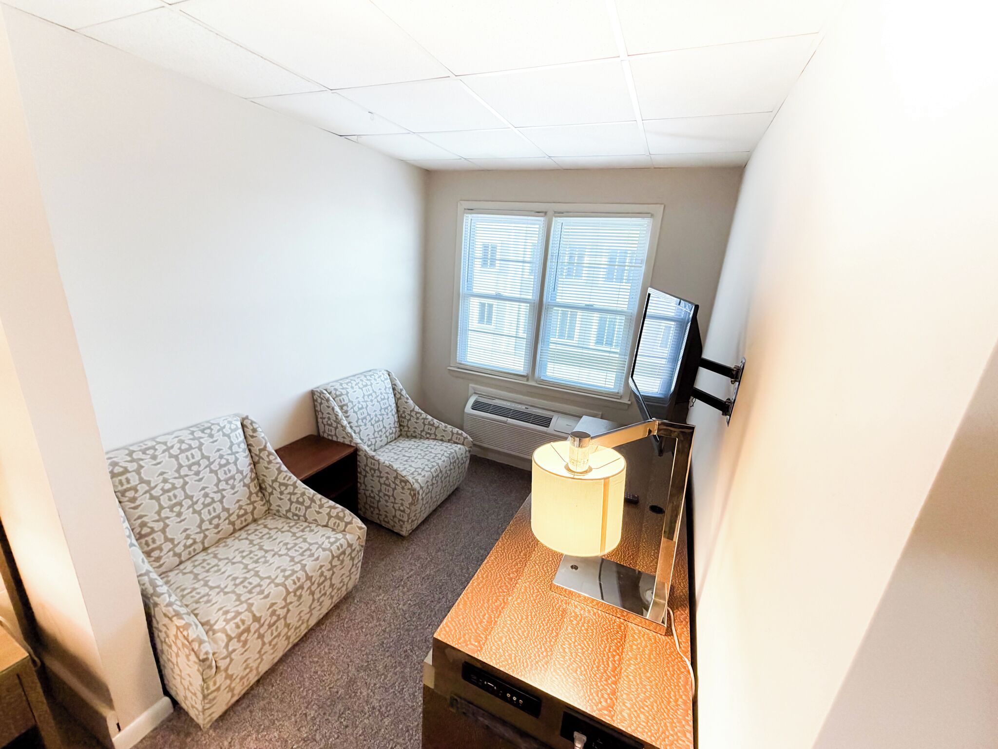 A small, well-lit room with a patterned sofa, a side table, a lamp, and a window with blinds.