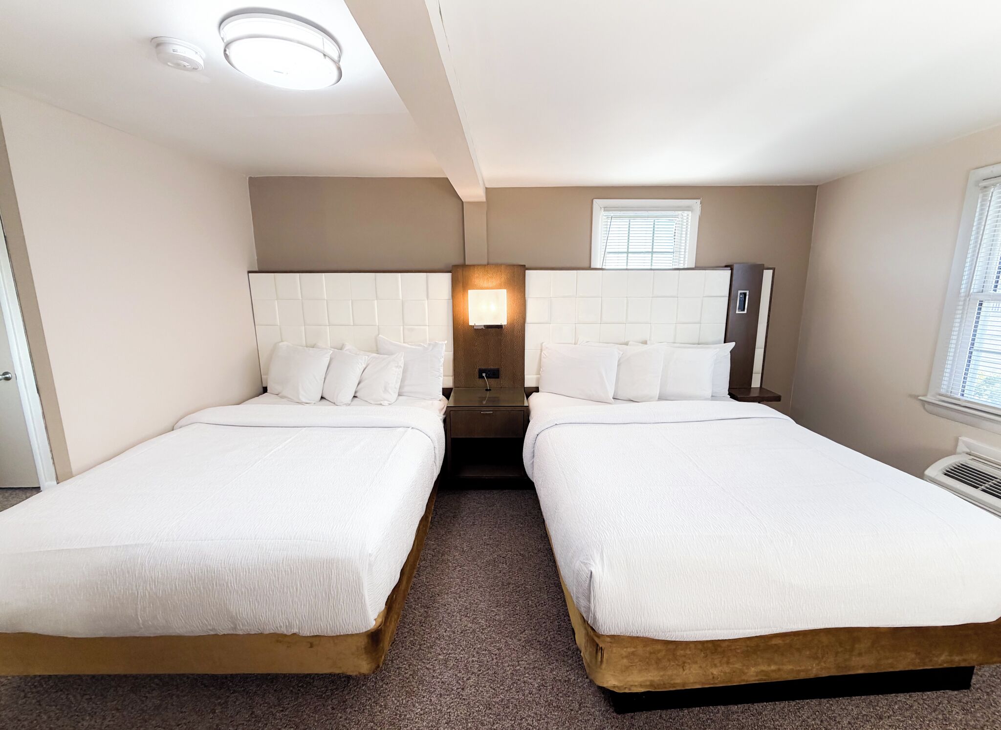 A hotel room with two beds, a nightstand, a wall-mounted lamp, and a window with blinds.