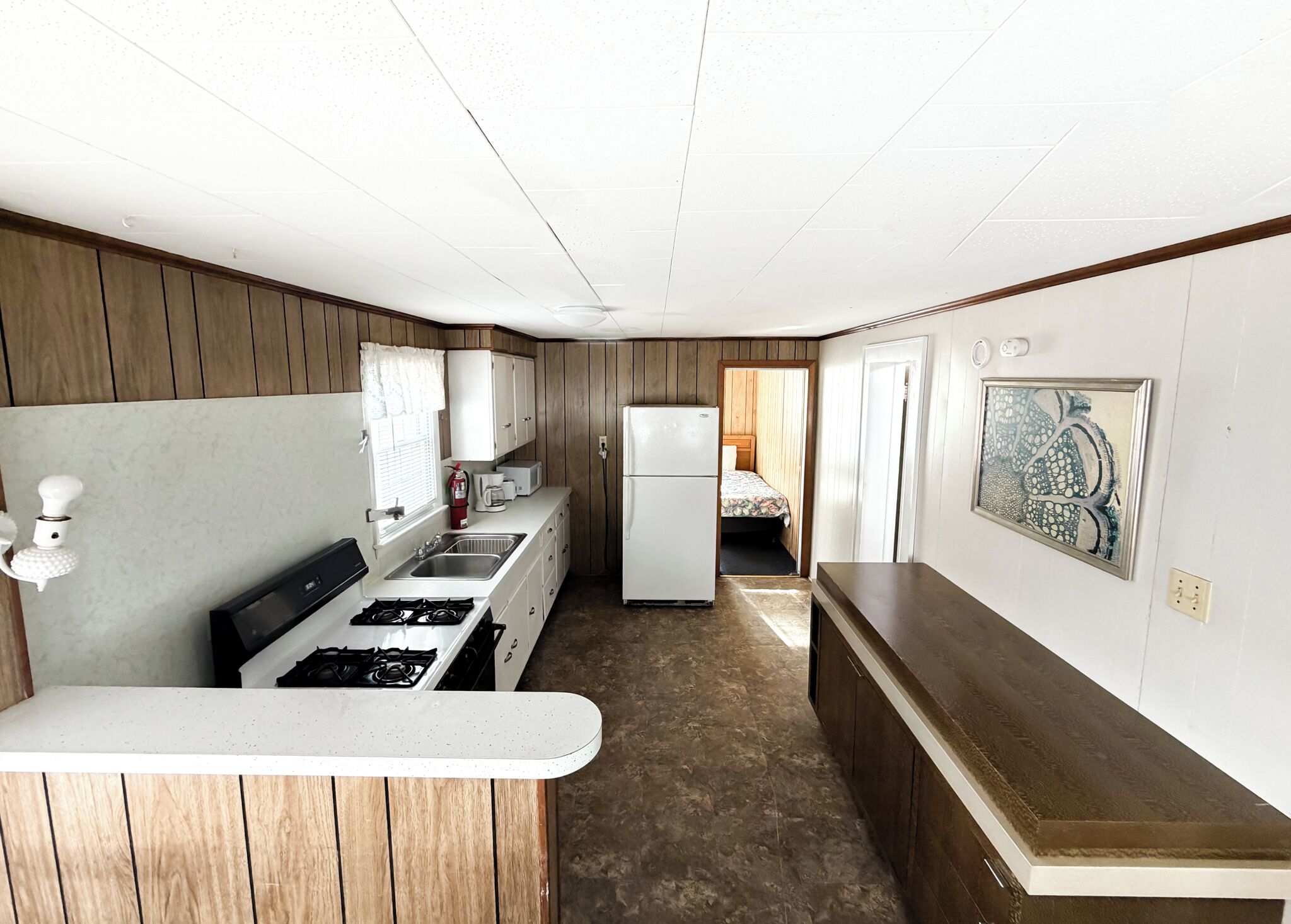 Una cocina compacta con estufa, lavamanos y refrigerador, paneles de madera y un pequeño dormitorio con cama y ventana.