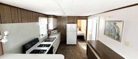 Una cocina compacta con estufa, lavamanos y refrigerador, paneles de madera y un pequeño dormitorio con cama y ventana.