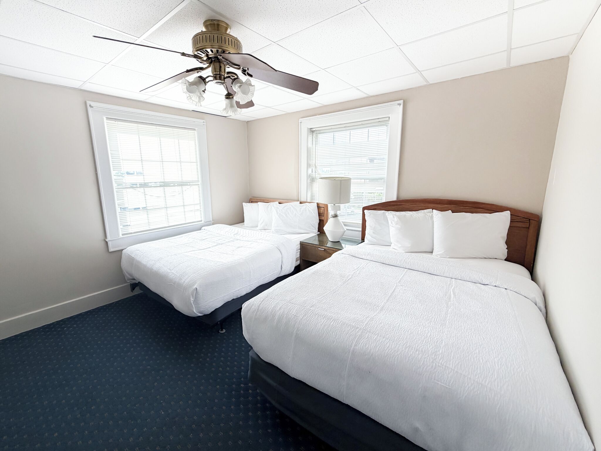 A hotel room with two beds, a ceiling fan, and large windows.