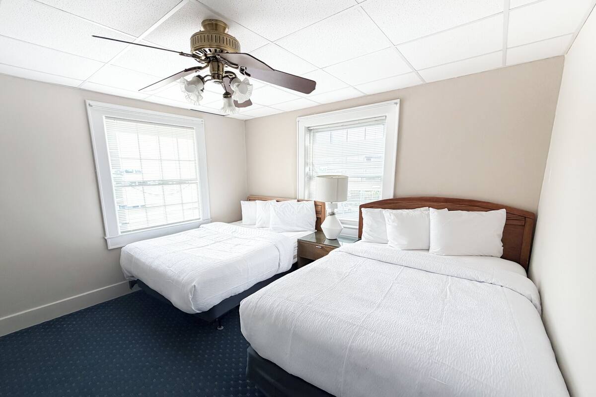Habitación de hotel con dos camas, ventilador de techo y ventanas amplias.