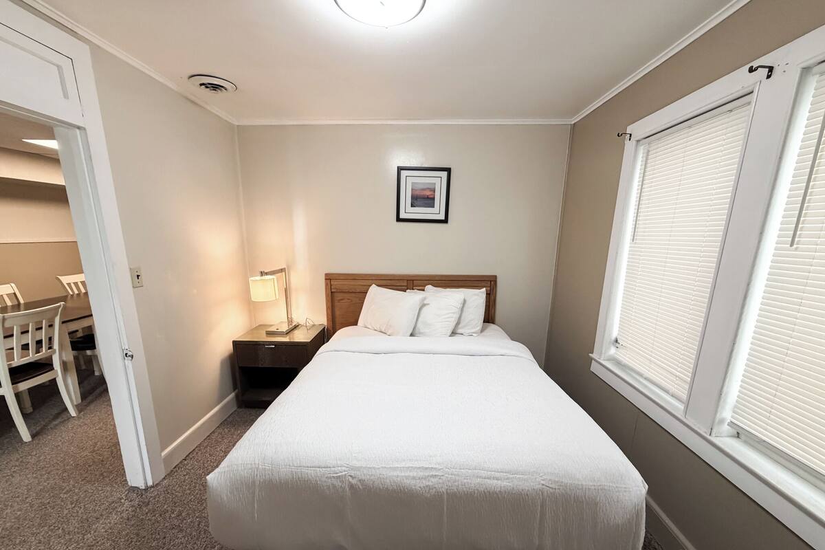 Un dormitorio con cama, mesita de noche, lámpara y ventana con persianas.