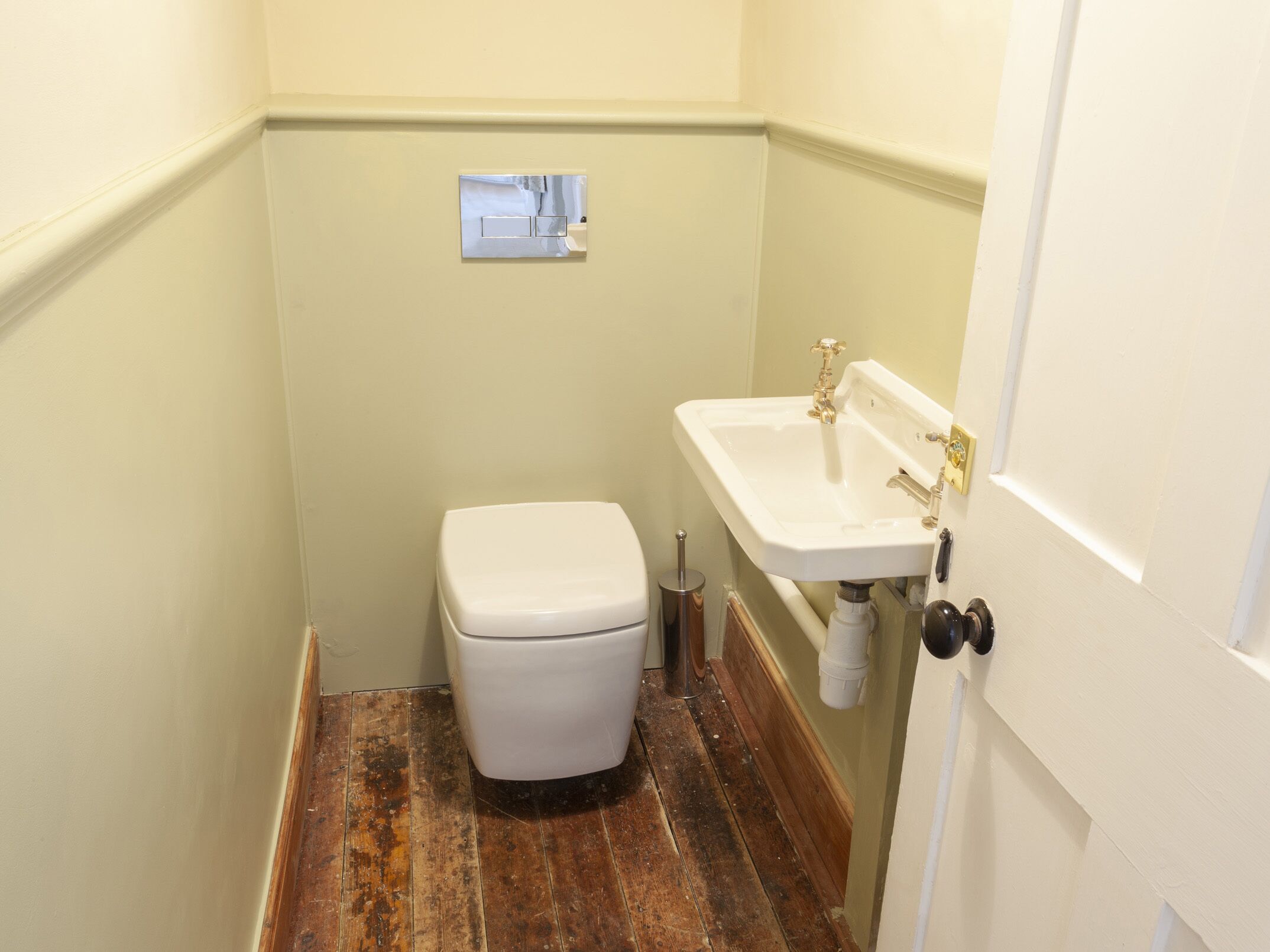 A small, narrow bathroom with a white toilet, a white sink with a gold faucet, and a wooden floor.