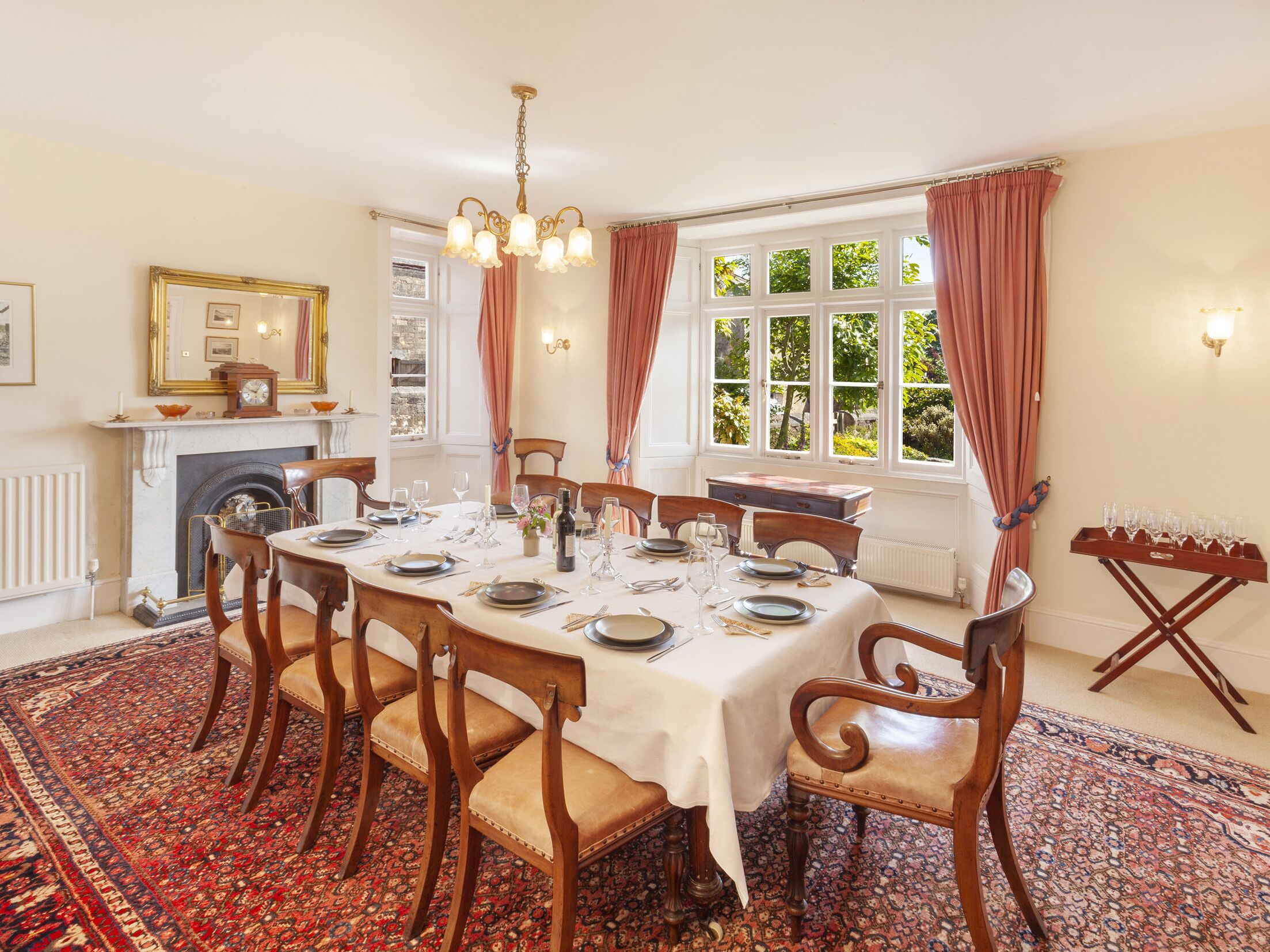 A dining room with a round table set for six, surrounded by wooden chairs with cushioned seats. A fireplace with a mantel is visible to the left, and a window with curtains is on the right.