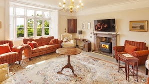 A spacious living room with a fireplace, comfortable seating, and a large window offering a view of greenery. - Crablake Farmhouse (Exeter)