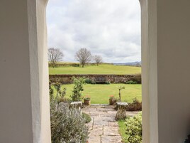 A stone pathway leading to a bench in a garden with a stone wall, flowering plants, and a grassy field with trees in the background.