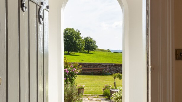 An arched doorway leading to a garden with a stone pathway, a brick wall, and a view of a green field and trees. - Crablake Farmhouse (Exeter)