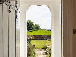 An arched doorway leading to a garden with a stone pathway, a brick wall, and a view of a green field and trees.