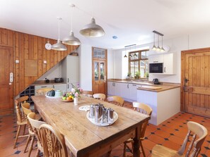 A kitchen with wooden cabinets, a dining table with chairs, hanging pendant lights, and a window with plants.