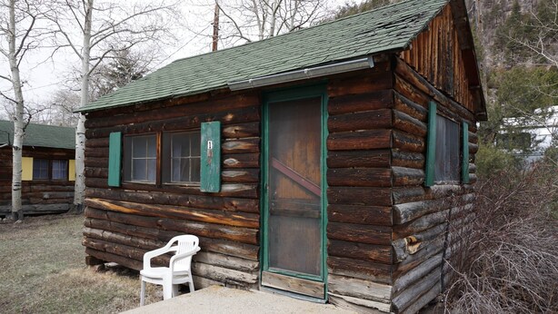 A rustic log cabin with a green door and windows, a concrete bench, and a chair outside.