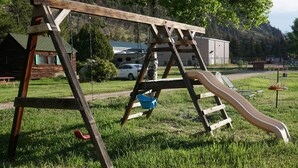 Un ensemble de jeux d’extérieur en bois, comprenant une balançoire et un toboggan, installé dans un espace herbeux, avec une cabane et des camping-cars en arrière-plan.