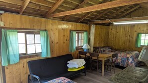 Une cabane chaleureuse aux murs en bois, avec un canapé, un lit recouvert d’une couverture colorée, une table à manger avec des chaises et une petite cuisine équipée d’une cuisinière.