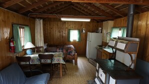 Un chalet confortable avec un intérieur en bois, un coin repas avec une table et des chaises, une petite cuisine équipée d’une cuisinière et un lit recouvert d’une couverture colorée.