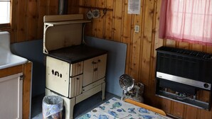 A cabin with a bed, a small kitchenette, a heater, and a window with a red and white checkered curtain.