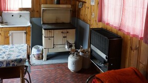 A rustic cabin interior with a wooden ceiling, a kitchenette with a stove, a bed with blankets, and a window with a red checkered curtain.
