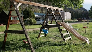 A wooden swing set with a slide in a grassy area with a cabin and RVs in the background.