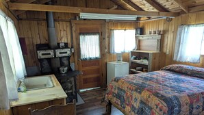 Une cabane avec un intérieur en bois, un lit recouvert d’une couverture aux couleurs vives, une petite cuisine équipée d’une cuisinière et une fenêtre avec des rideaux.