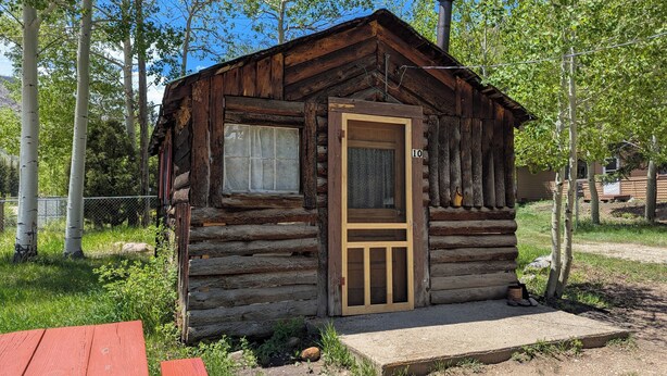 Un chalet rustique en rondins de bois, doté d’une porte et d’une fenêtre en bois, entouré d’arbres et agrémenté d’une table de pique-nique.