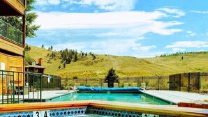 A swimming pool with wooden deck and metal railings, surrounded by a fenced area with a view of a hilly landscape. - Walkout Studio Near W RMNP + Pool & Hot Tubs (Granby)