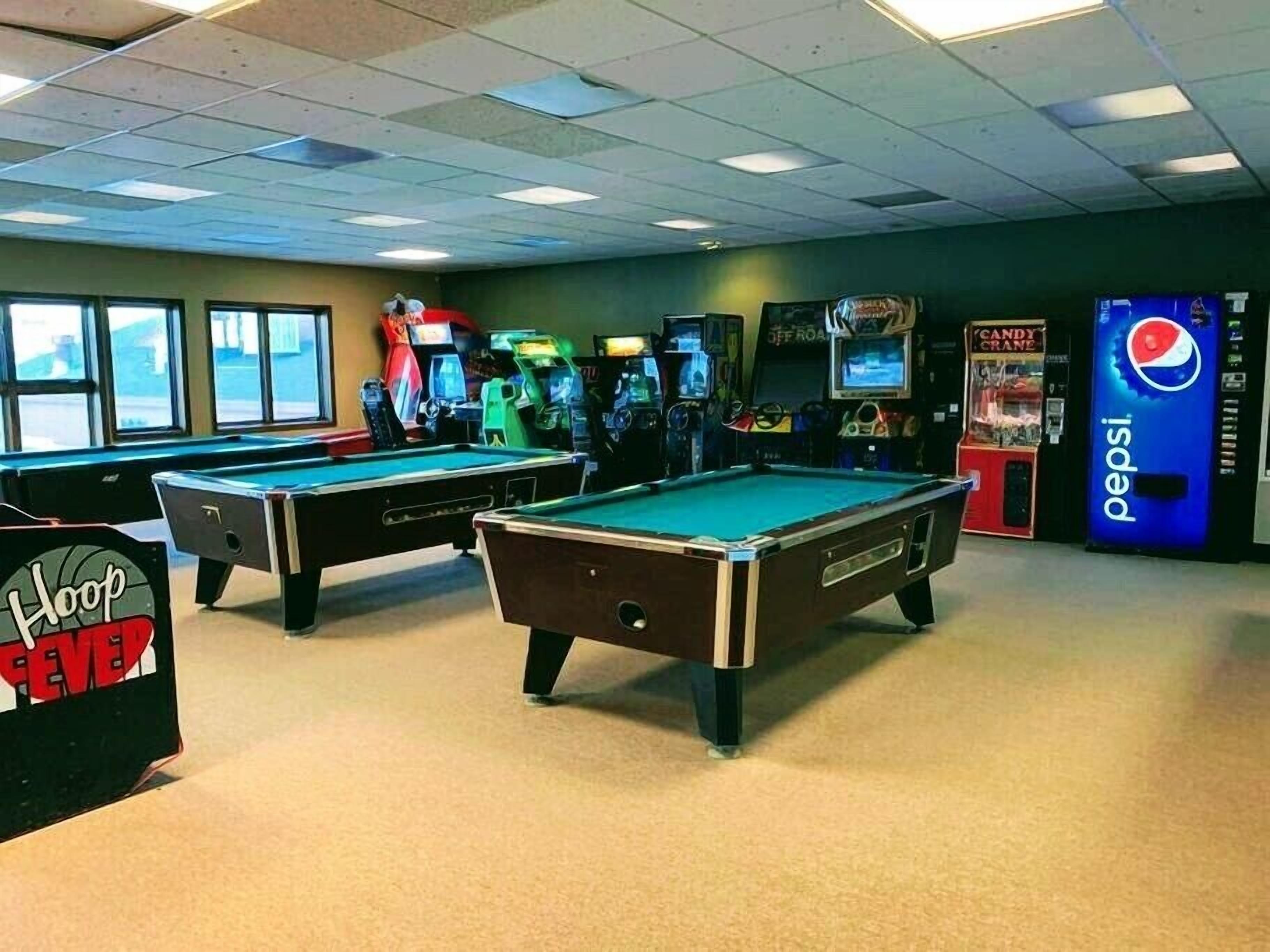 Une salle de billard avec deux tables de billard, des jeux d’arcade et un distributeur automatique de boissons Pepsi.