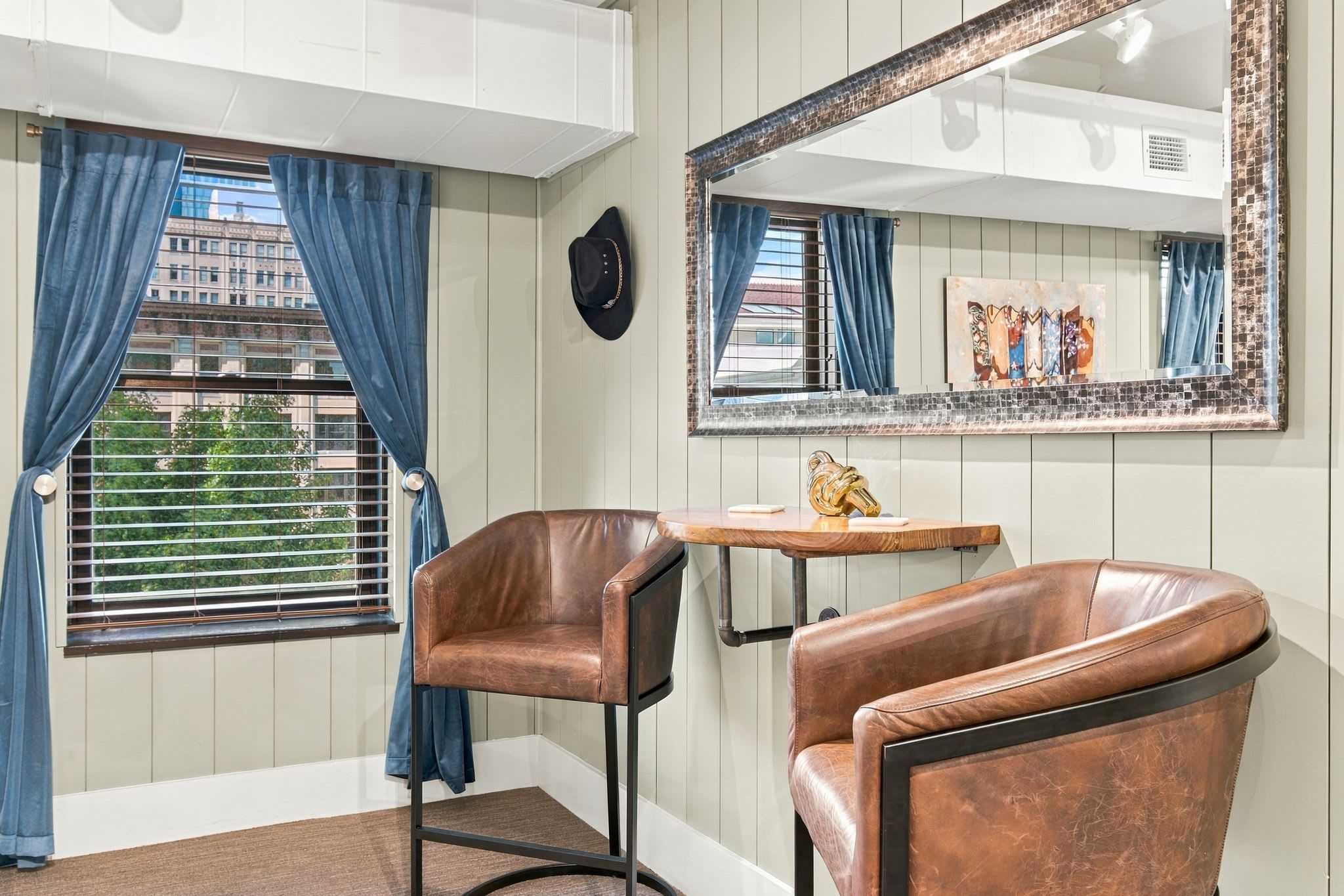 A room with two leather chairs, a small wooden table, a mirror, and a window with blue curtains.