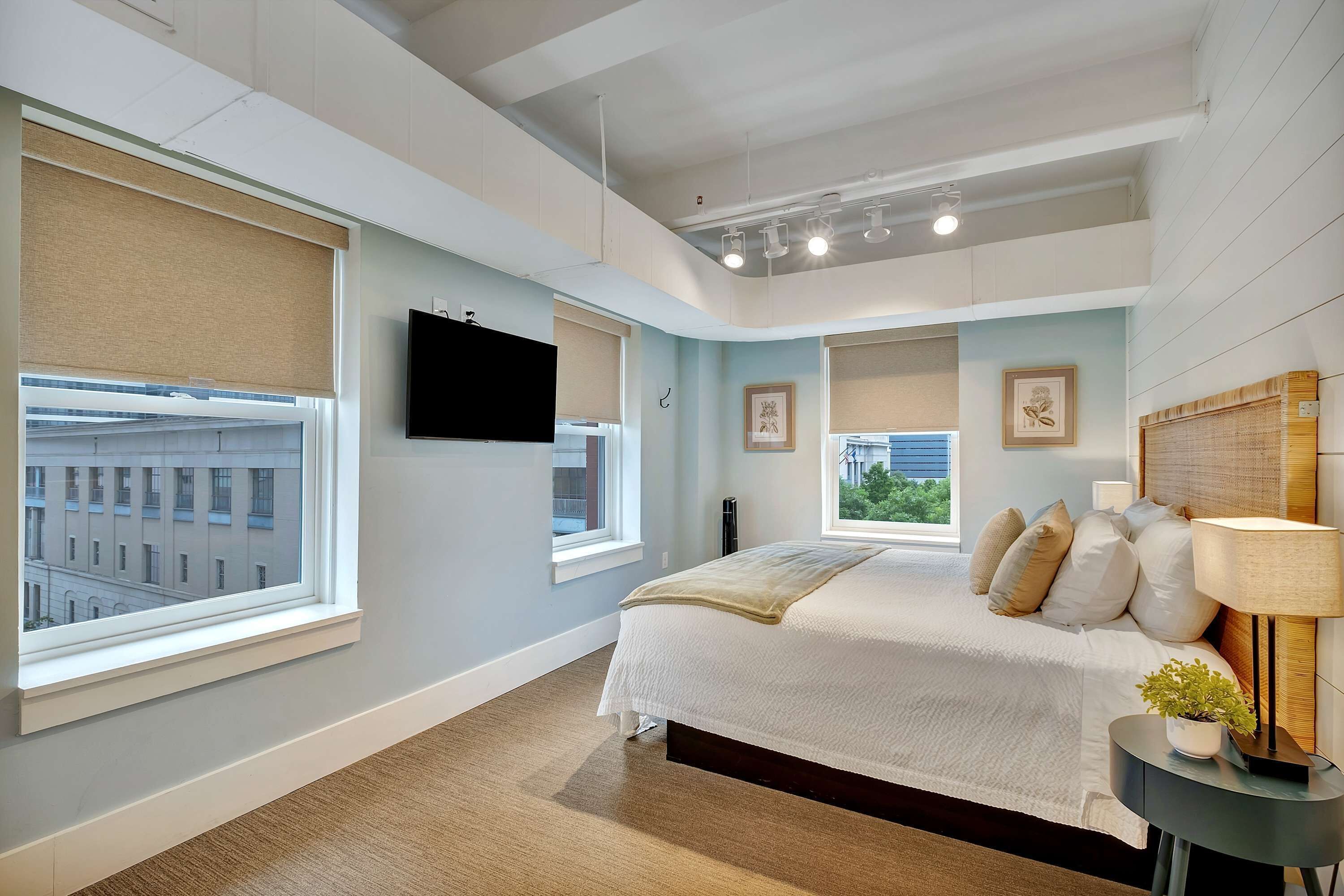 A bedroom with a bed, a TV, a bedside table with a lamp, and windows with blinds.