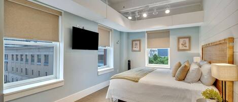 A bedroom with a bed, a TV, a bedside table with a lamp, and windows with blinds.