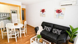 Ein modernes Wohnzimmer mit einer schwarzen Ledersofa, einem weißen Esstisch und Stühlen, einem Couchtisch mit einer Pflanze und einer Küche im Hintergrund.