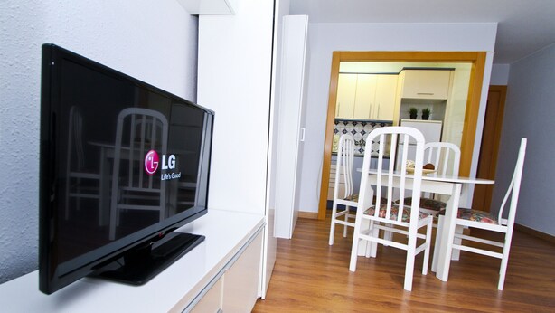 Ein modernes Wohnzimmer mit einem Flachbildfernseher auf einem weißen Schrank, einem Essbereich mit weißen Stühlen und einer durch eine offene Tür einsehbaren Küche.