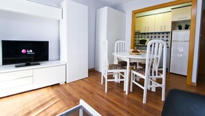 Ein kompakter Wohnbereich mit Flachbildfernseher auf einer weißen Unterhaltungseinheit, einem Esstisch mit weißen Stühlen und einer Küchenzeile mit Schränken und einer Mikrowelle.