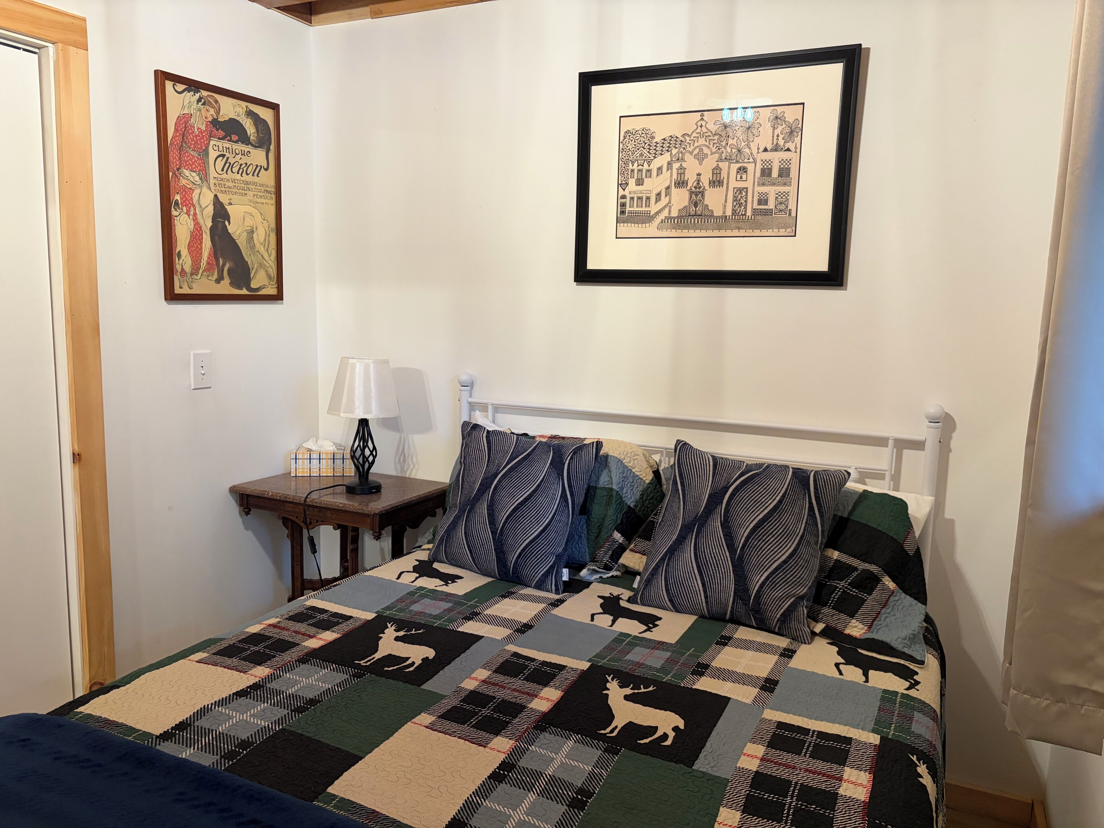 A bedroom with a bed, pillows, a bedside table with a lamp, a framed picture on the wall, and a quilt with animal patterns.
